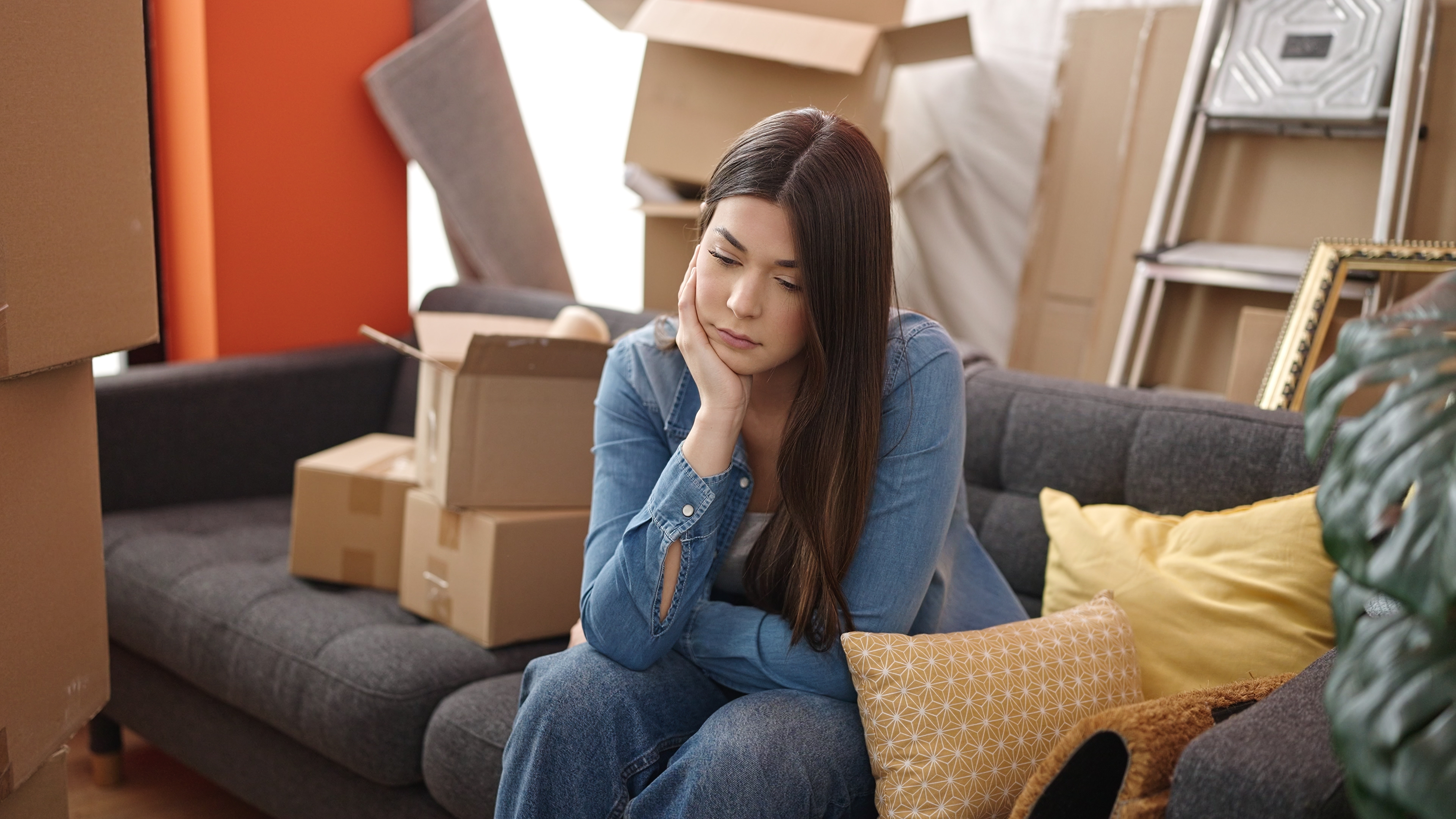 Persona sentada en un sofá rodeada de cajas de mudanza en un nuevo hogar, con expresión reflexiva, representando el proceso de adaptación asociado al duelo migratorio